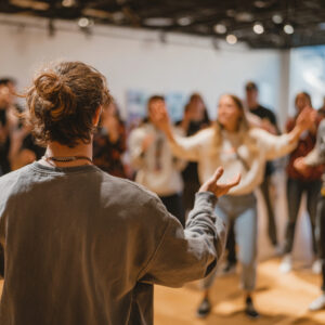 In a room with an exposed ceiling, people are playing the game Simon Says, with the back of a person playing as Simon and the a group of people looking at them and copying their actions.